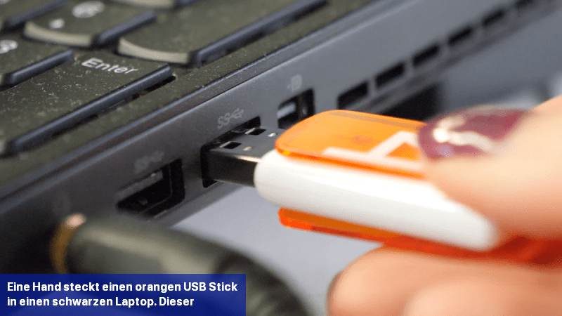 Eine Hand steckt einen orangen USB-Stick in einen schwarzen Laptop. Dieser wird gerade von einem Stromkabel geladen.
